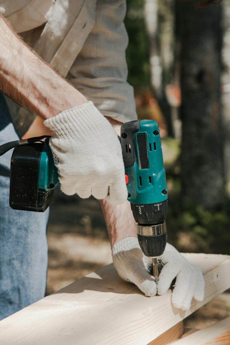 A construction worker using a cordless drill on a wooden panel outdoors, wearing protective gloves.